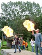 Zwei Männer auf einem Podest spucken jeweils eine große Flamme in die Luft. Im Hintergrund ist ein Spielgerät aus Holz mit einigen Kindern zu erkennen.