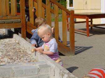 Zwei Kleinkinder spielen an einer niedrigen Betonmauer. Neben ihnen eine Holztreppe, im Hintergrund ein Holztisch.