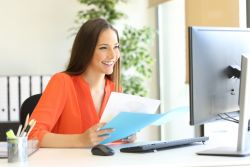 Eine lachende junge Frau in orangefarbener Bluse sitzt in einem modernen Büro am Schreibtisch und schaut auf den Computerbildschirm. In der Hand hält sie mehrere verschiedenfarbige Zettel.