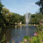 Kurpark Blick über einen Teich zwischen blühenden Pflanzen hindurch. Mitten im Teich eine Fontäne, an der sich ein Regenbogen bildet. Dahinter eine halbkreisförmige Bühne mit einem überstehenden Dach.
