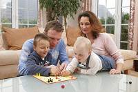 Mann, Frau und zwei kleine Kinder spielen in einem Wohnzimmer ein Brettspiel.