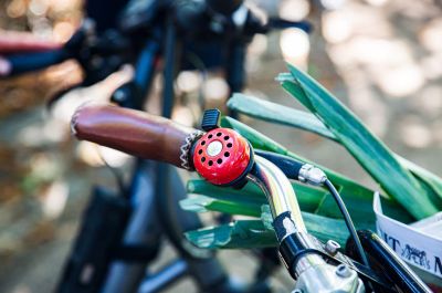 Ein Fahrradlenker mit einer auffälligen roten Klingel und einem Korb, der mit Gemüse gefüllt ist. Im unscharfen Hintergrund befindet sich weitere Fahrräder.