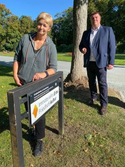 Eine Frau in einer gepflegten Grünanlage lehnt sich auf ein Schild mit der Aufschrift "Immaterielles Erbe Friedhofskultur".  Hinter ihr steht Oberbürgermeister Feist.