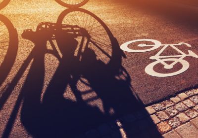 Im eng begrenzten Ausschnitt einer geteerten Oberfläche ist das Piktogramm eines Fahrrades dargestellt. Durch die sehr tief stehende Sonne wird ins den Ausschnitt hinein der lange Schatten eines Fahrrades geworfen, das außerhalb des Bildausschnittes steht.