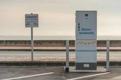 Rechteckige weiße Säule mit der Aufschrift "Stromtankstelle" an einem öffentlichen Parkplatz am Südstrand. Daneben ein Schild "Ladeplatz - Nur für Elektrofahrzeuge". 