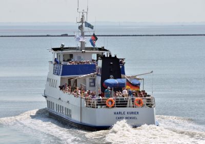 Das vollbesetzte Ausflugsschiff Harlekurier fährt an einem sonnigen Tag hinaus aufs Meer. Das Schiff ist von hinten zu sehen und hinterlässt durch die hohe Geschwindigkeit eine Spur aus aufgewühltem Wasser.