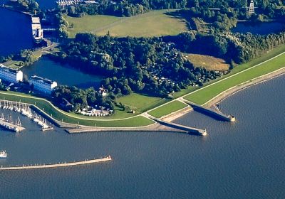 Luftbildaufnahme aus größerer Höhe. Zwischen dem Meer am unteren Bildrand und dem Innenhafen oben liegt eine Landfläche aus Grasflächen und Baumbeständen. Zwei parallele Molen ziehen sich vom Meer durch den Landbereich in den inneren Hafen.