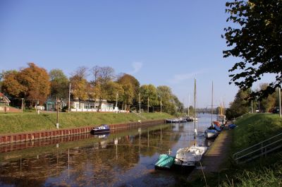 Herbststimmung am Rüstersieler Hafen mit einigen Segelbooten. Auf der anderen Uferseite hinter der rostfarbenen Spundwand ist ein großes Gebäude mit Biergarten zu erkennen. Im weiteren Verlauf des Gewässers ist am Horizont eine Brücke zu erkennen.