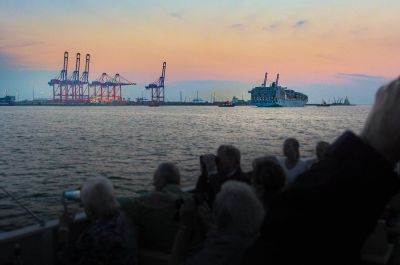 Brücken des Container-Terminals im Sonnenuntergang. Davor ein Containerschiff. Das Foto wurde über die Köpfe zahlreicher Menschen an Bord eines Schiffes aufgenommen, von denen einige mit dem Smartphone fotografieren oder durch ein Fernglas blicken.