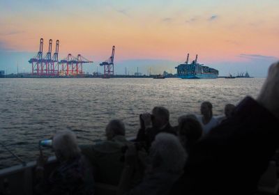 Brücken des Container-Terminals im Sonnenuntergang. Davor ein Containerschiff. Das Foto wurde über die Köpfe zahlreicher Menschen an Bord eines Schiffes aufgenommen, von denen einige mit dem Smartphone fotografieren oder durch ein Fernglas blicken.
