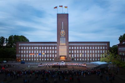 Frontalansicht des Rathauses in der Abenddämmerung. Vor dem Rathaus stehen in einer Reihe uniformierte Musiker und Matrosen mit Fackeln in der Hand. Auf dem Rathausplatz stehen zahlreiche Menschen.