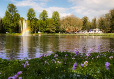 Ein Teich mit einer Wasserfontäne, an der sich ein Regenbogen bildet. Im Vordergrund eine Rasenfläche mit Frühjahrsblumen. Im Hintergrund ein Gebäude mit einem Biergarten.