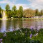 Ein Teich mit einer Wasserfontäne, an der sich ein Regenbogen bildet. Im Vordergrund eine Rasenfläche mit Frühjahrsblumen. Im Hintergrund ein Gebäude mit einem Biergarten.