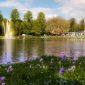 Ein Teich mit einer Wasserfontäne, an der sich ein Regenbogen bildet. Im Vordergrund eine Rasenfläche mit Frühjahrsblumen. Im Hintergrund ein Gebäude mit einem Biergarten.