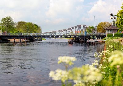 Deichbrücke an einem sommerlichen Tag. Aufgenommen aus Richtung des Großen Hafens über die Blüten der Pflanzen an der Uferböschung.